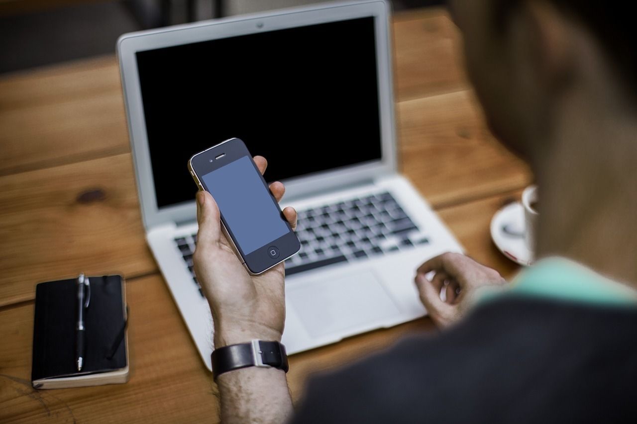 A man holding a mobile phone, sitting in front of a laptop.