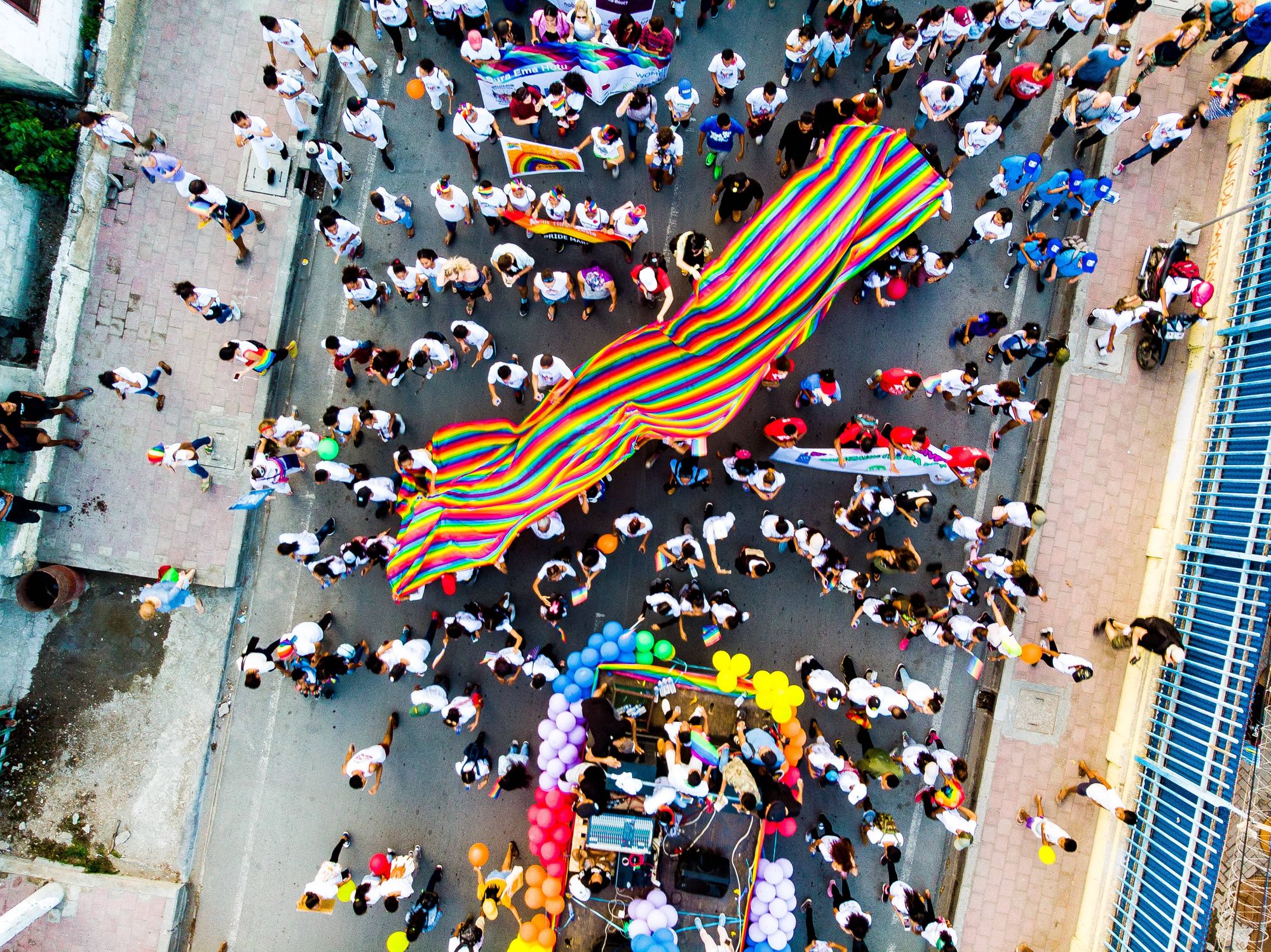 A birds-eye-view of a street with a Pride rally happening below. People are holding up rainbow flags and coloured balloons.