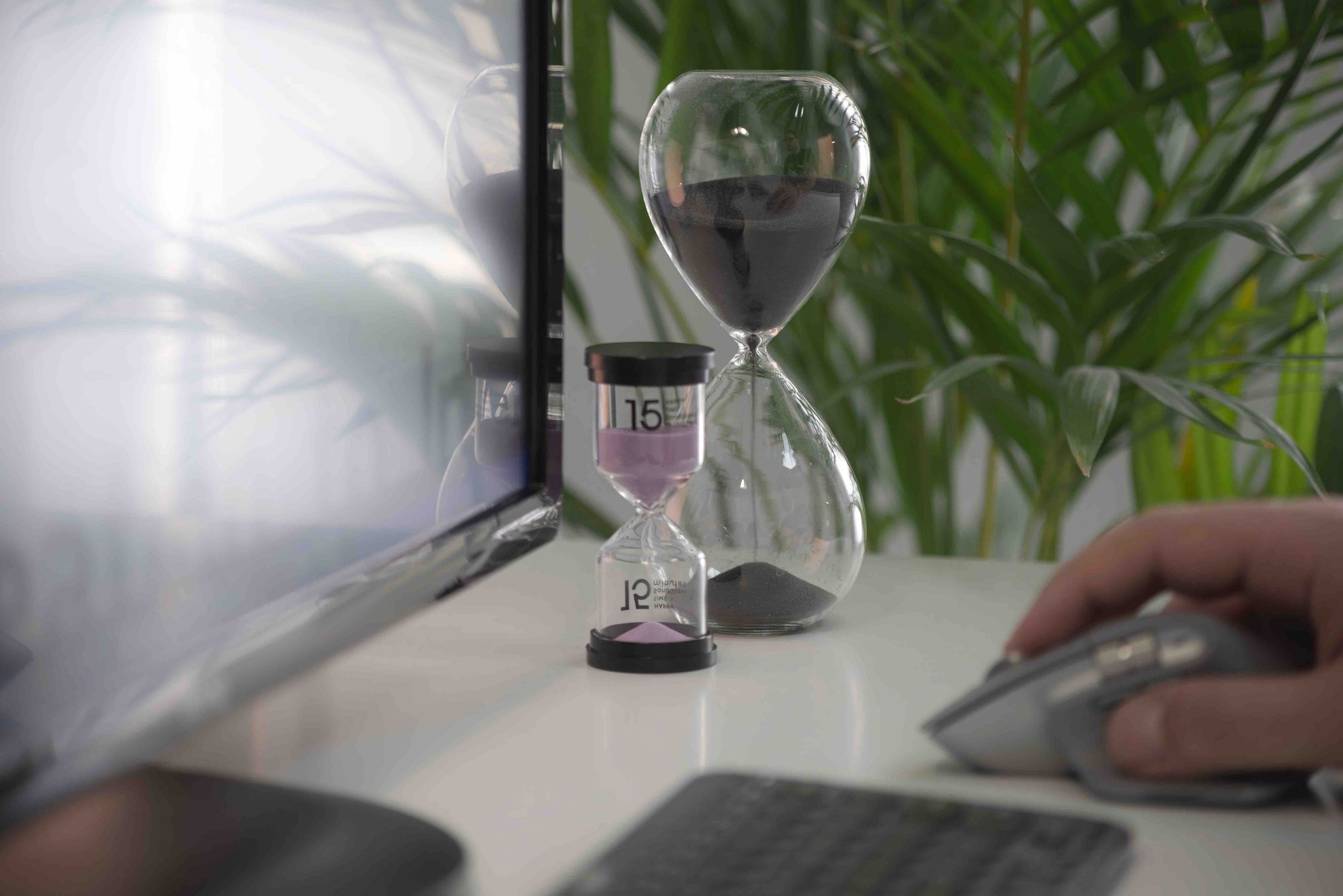 Two hourglasses sat on a desk, a hand is coming into shot from the right hand side.