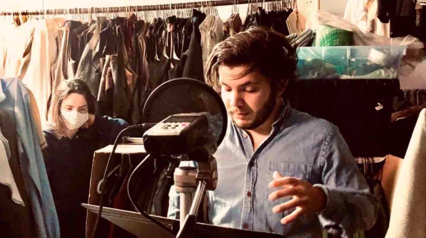 A man recording a podcast in a room filled with clothes.